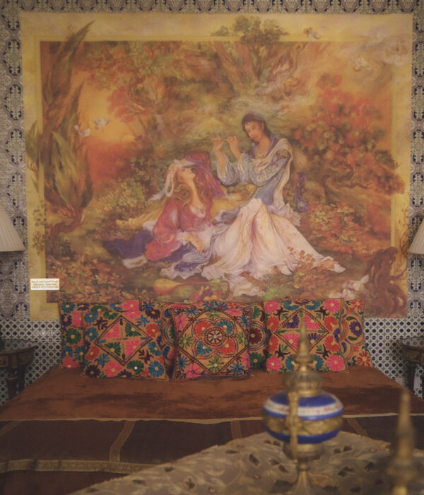 Bedroom corner with large painting, embroidered cushions, wooden seating, and arabesque patterned walls.