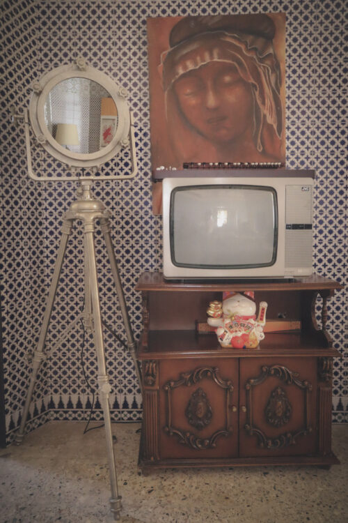 Vintage CRT TV on wooden cabinet with patterned tiles, mirror on tripod, and wall painting above.