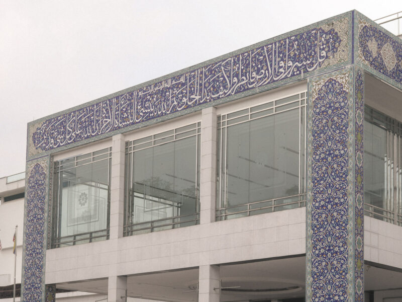 Exterior of the Islamic Arts Museum in Kuala Lumpur, showing blue Islamic tilework, Arabic calligraphy, glass windows, and modern white architecture.