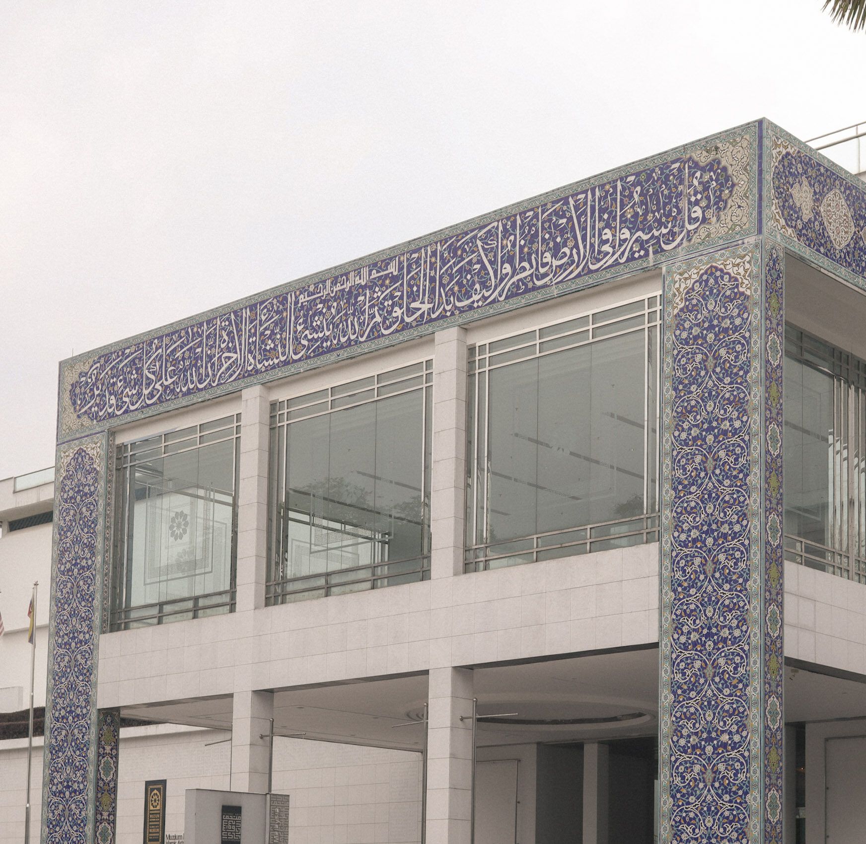 Exterior of the Islamic Arts Museum in Kuala Lumpur, showing blue Islamic tilework, Arabic calligraphy, glass windows, and modern white architecture.