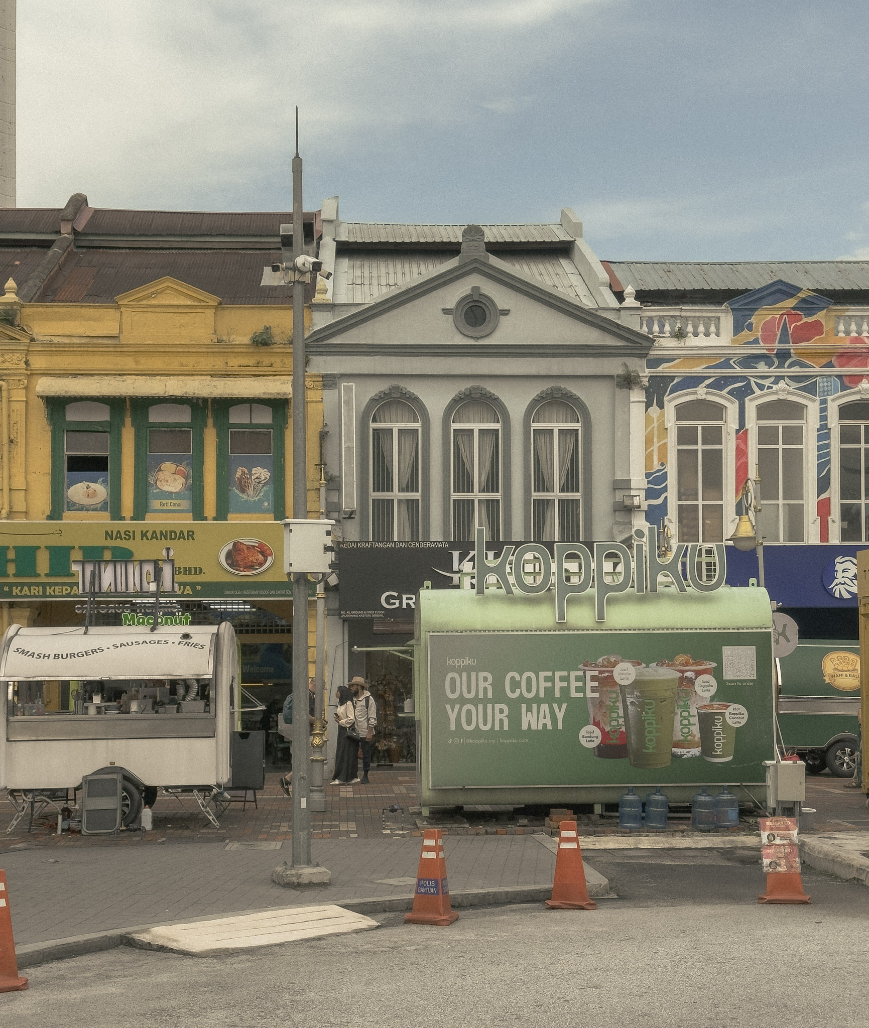 Street view near Central Market in Kuala Lumpur, showing colorful old shophouse facades, food signs, a coffee kiosk, street cones, and people walking on the sidewalk.