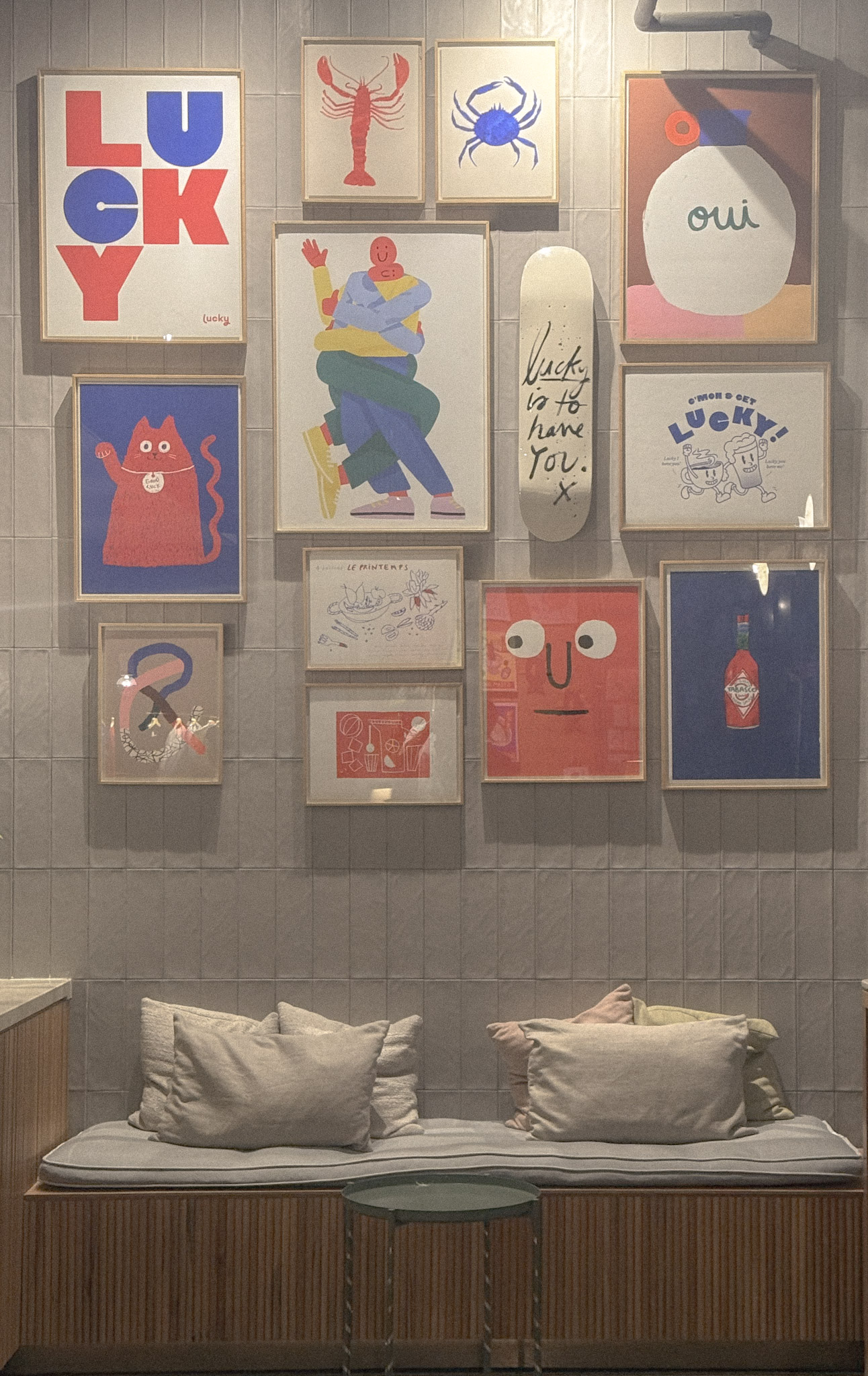 A wall display of colorful framed illustrations and posters, including the word “LUCKY,” a red cat, a lobster, a crab, and abstract figures, above a cushioned bench with pillows.