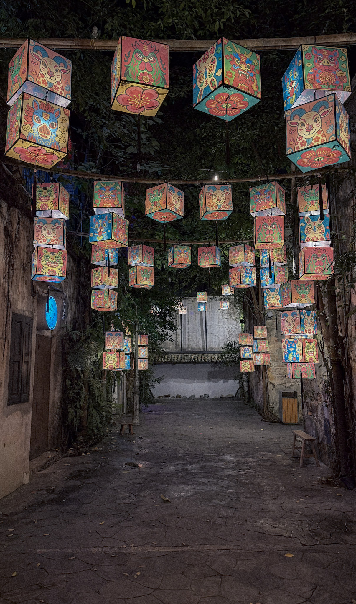 Colorful illustrated lanterns hanging above a narrow walkway at Kwai Chai Hong in Kuala Lumpur, with old walls, trees, benches, and a dim evening atmosphere.