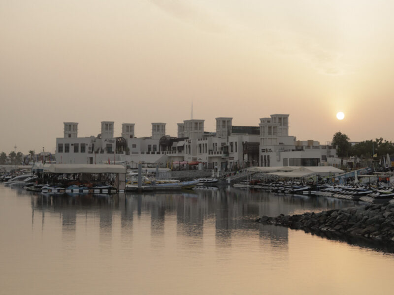 A calm UAE harbor at sunrise with traditional-style buildings reflected on the water