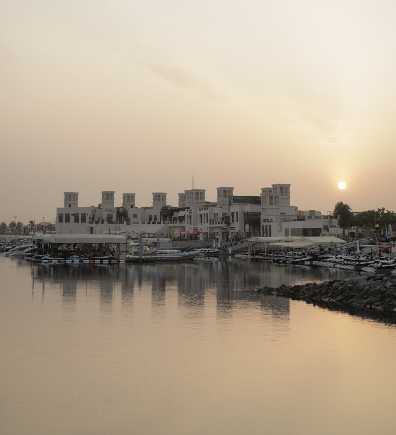 A calm UAE harbor at sunrise with traditional-style buildings reflected on the water